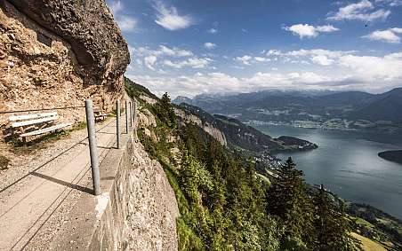 Blick auf einen Bergpfad mit Bänken und See im Hintergrund.