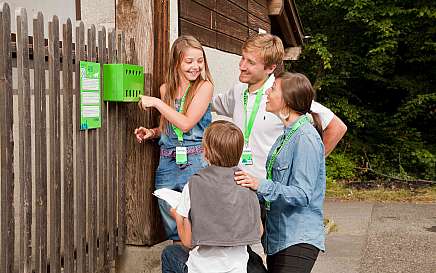 Eine Familie interagiert mit einem grünen Informationskasten an einem Zaun.