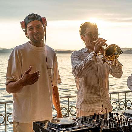A DJ and a trumpeter play music at sunset by the water.