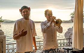 Ein DJ und ein Trompeter spielen Musik bei Sonnenuntergang am Wasser.