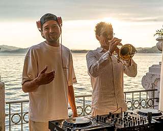 Ein DJ und ein Trompeter spielen Musik bei Sonnenuntergang am Wasser.