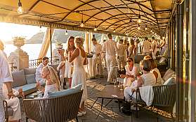 Menschen in weißen Outfits genießen eine Feier auf einer Terrasse am Wasser.