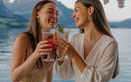 Zwei Frauen stoßen mit Getränken an, umgeben von Blumen und Wasser im Hintergrund