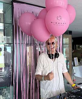 A DJ stands with pink balloons and a mixing console.