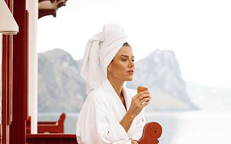 Woman in bathrobe enjoying croissant on a balcony with mountain view.