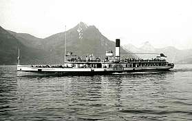 Ein Dampfschiff fährt auf einem ruhigen See mit Bergen im Hintergrund.