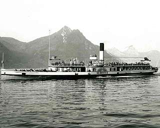 Ein Dampfschiff fährt auf einem ruhigen See mit Bergen im Hintergrund.