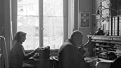 Eine Frau tippt an einer Schreibmaschine, ein Mann telefoniert.