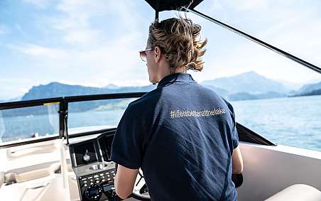 Junge Person steuert ein Boot am See mit Bergen im Hintergrund.