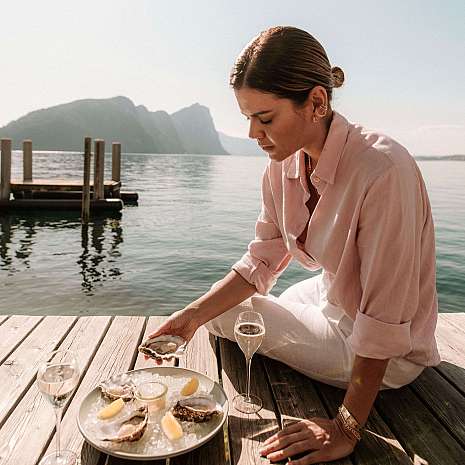 A woman sits by the water enjoying oysters with champagne.