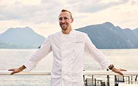 Ein Koch in weißer Uniform steht am Ufer mit Bergen im Hintergrund.
