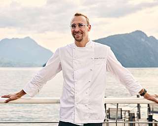 Ein Koch in weißer Uniform steht am Ufer mit Bergen im Hintergrund.