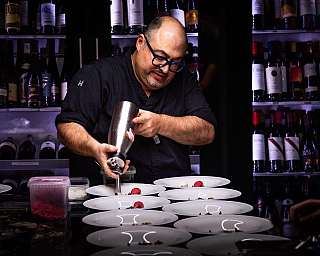 Chef using a whipped cream dispenser to plate desserts in a kitchen with wine bottles