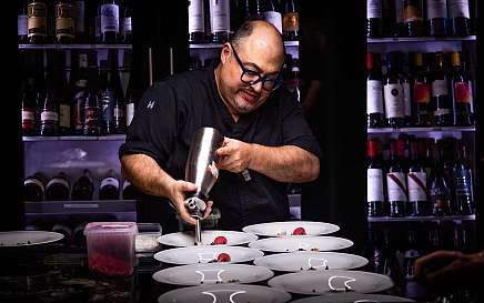 Chef using a whipped cream dispenser to plate desserts in a kitchen with wine bottles
