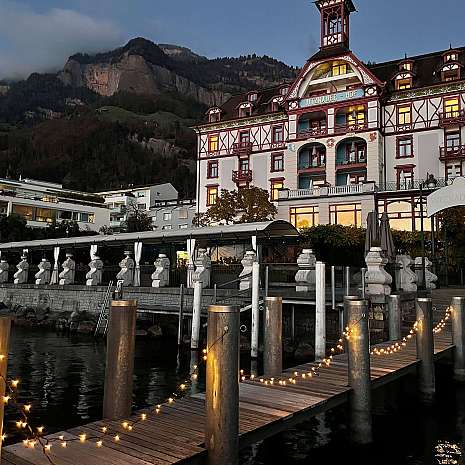 Abendaufnahme eines beleuchteten Stegs vor einem historischen Hotel mit Bergen im Hintergrund