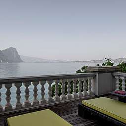 Blick von einer Terrasse auf einen ruhigen See mit Bergen im Hintergrund.