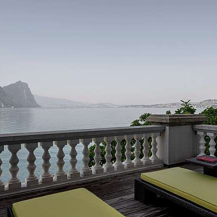 Blick von einer Terrasse auf einen ruhigen See mit Bergen im Hintergrund.